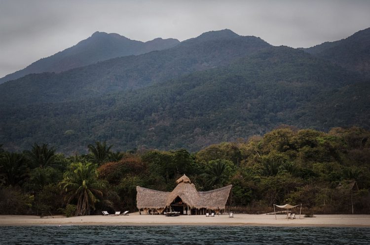 „digitale Detox“-rhino-africa-travel-remote-places-greystoke mahale hut on lake beach in Tanzania Das Meer, dahinter Strand mti einem Haus, dahinter grüne Berge