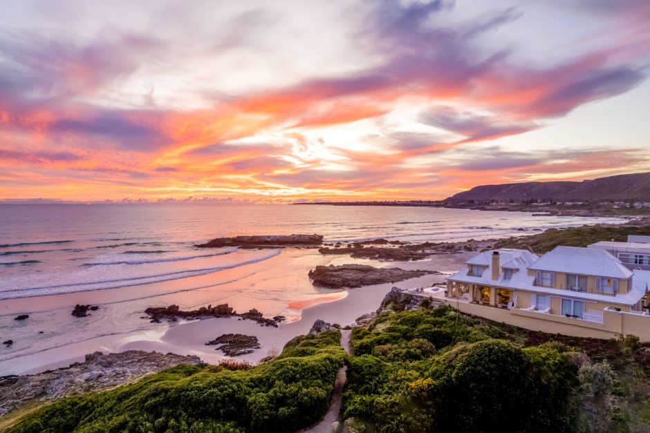 Birkenhead House overlooks the ocean and a rocky beach in Hermanus, with a colourful sunset lighting up the sky