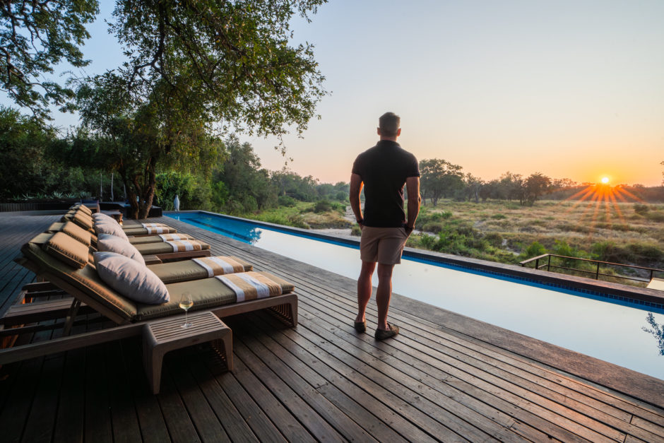 A man in a black polo shirt and khaki shorts stands on the Jackalberry breakfast deck at Silvan Safari lodge, gazing out over the Manyeleti Riverbed at sunrise. An infinity pool and lined-up sun loungers occupy the wooden deck, with a lone glass of wine resting on a small table. Trees and bushland stretch into the distance under the warm morning light.