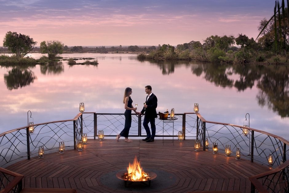 A couple enjoys an intimate moment on a lantern-lit wooden deck with a crackling fire, overlooking a calm river under a pastel sunset sky