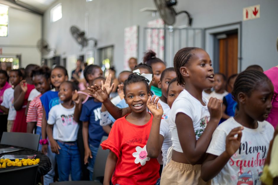 A group of children stand in a lively queue, some waving and smiling brightly, while others chat and laugh, filling the space with energy and excitement.