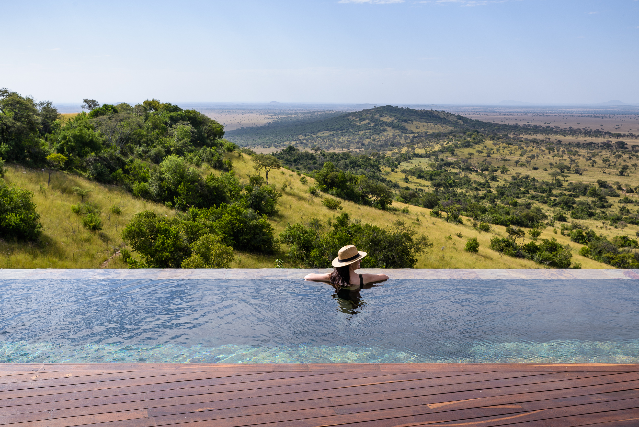 A woman in a straw hat relaxes in an infinity pool overlooking the African savannah with rolling hills and open plains.