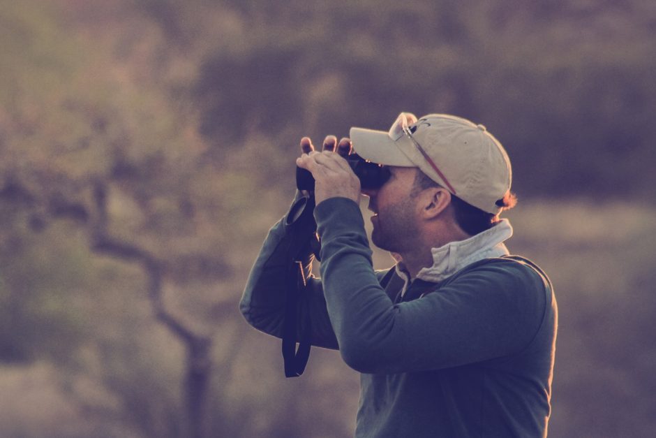 David wearing a baseball cap and jacket peers intently through binoculars in a natural setting with blurred trees in the background