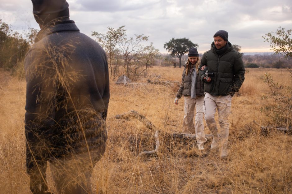 Atlas&Arc-RhinoAfrica-5283 A couple dressed in warm outdoor clothing walks arm-in-arm through dry grassland, following a guide during a guided nature walk in an African bush landscape under a cloudy sky.