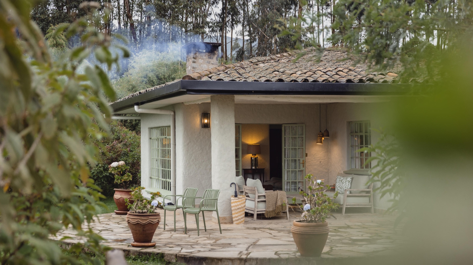 Sabyinyo-Silverback-Lodge-suite A cosy cottage with soft lighting, outdoor seating, and smoke rising from the chimney is surrounded by potted plants and leafy trees.