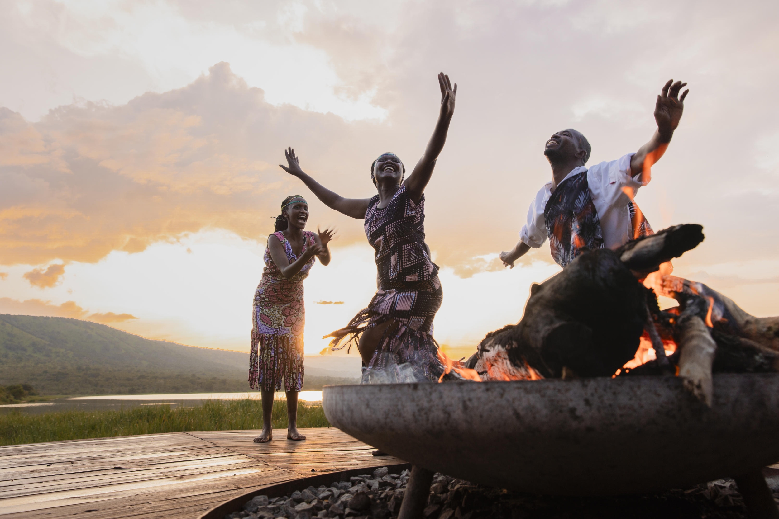 Wilderness Magashi Three barefoot dancers perform around a glowing firepit at sunset, their hands raised in joy against a lake and mountain backdrop.