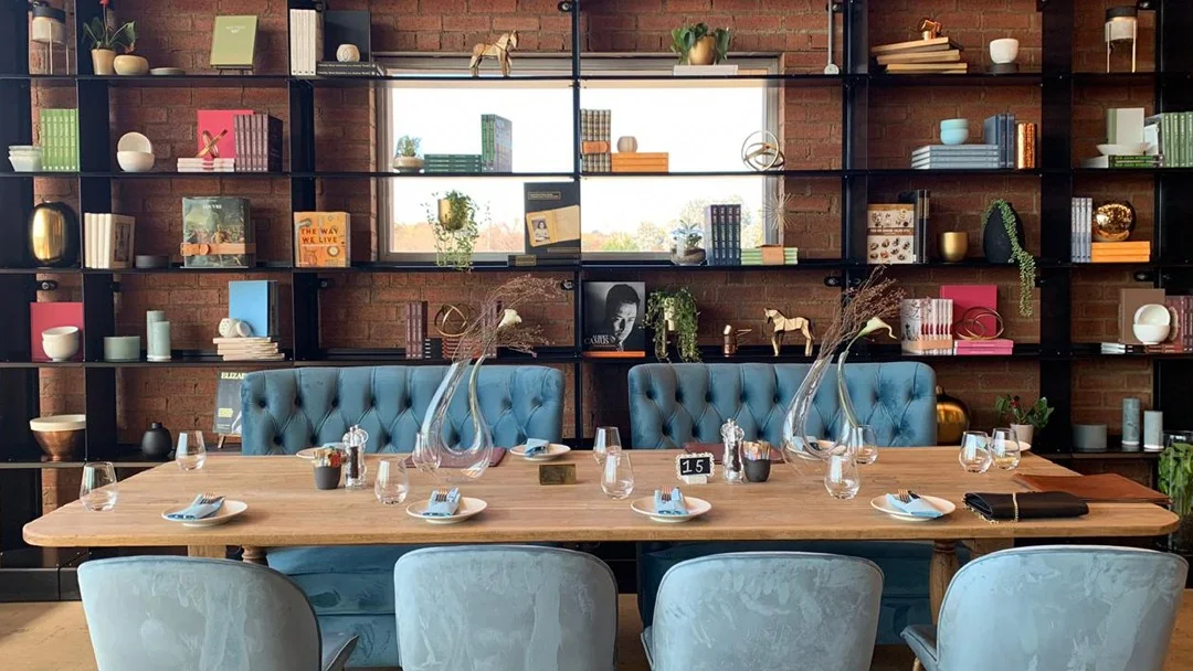 olives-and-plates-hyde-park A long dining table with place settings and blue couches and chairs around it and a bookcase behind it