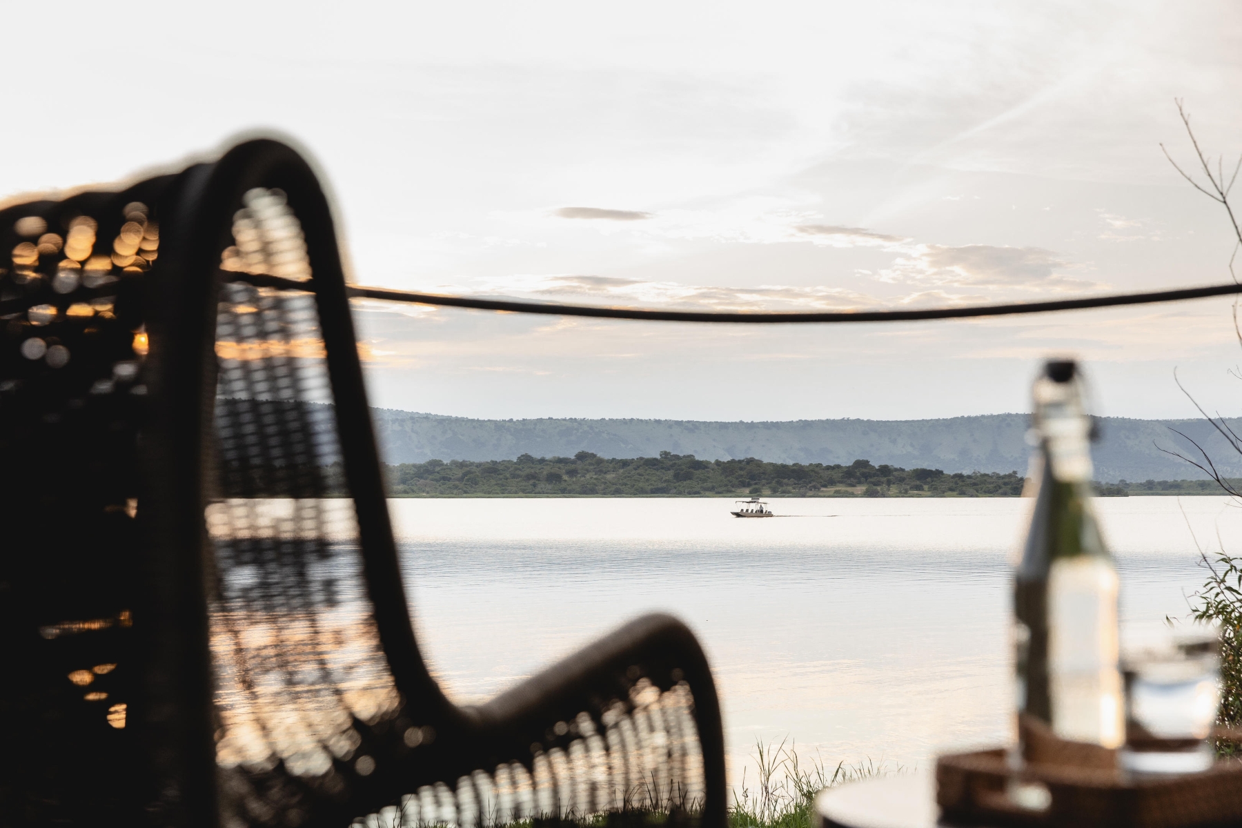 Wilderness-Magashi-view A boat glides across a still lake at sunset, viewed through the frame of a wicker chair and a table with a bottle and glass.
