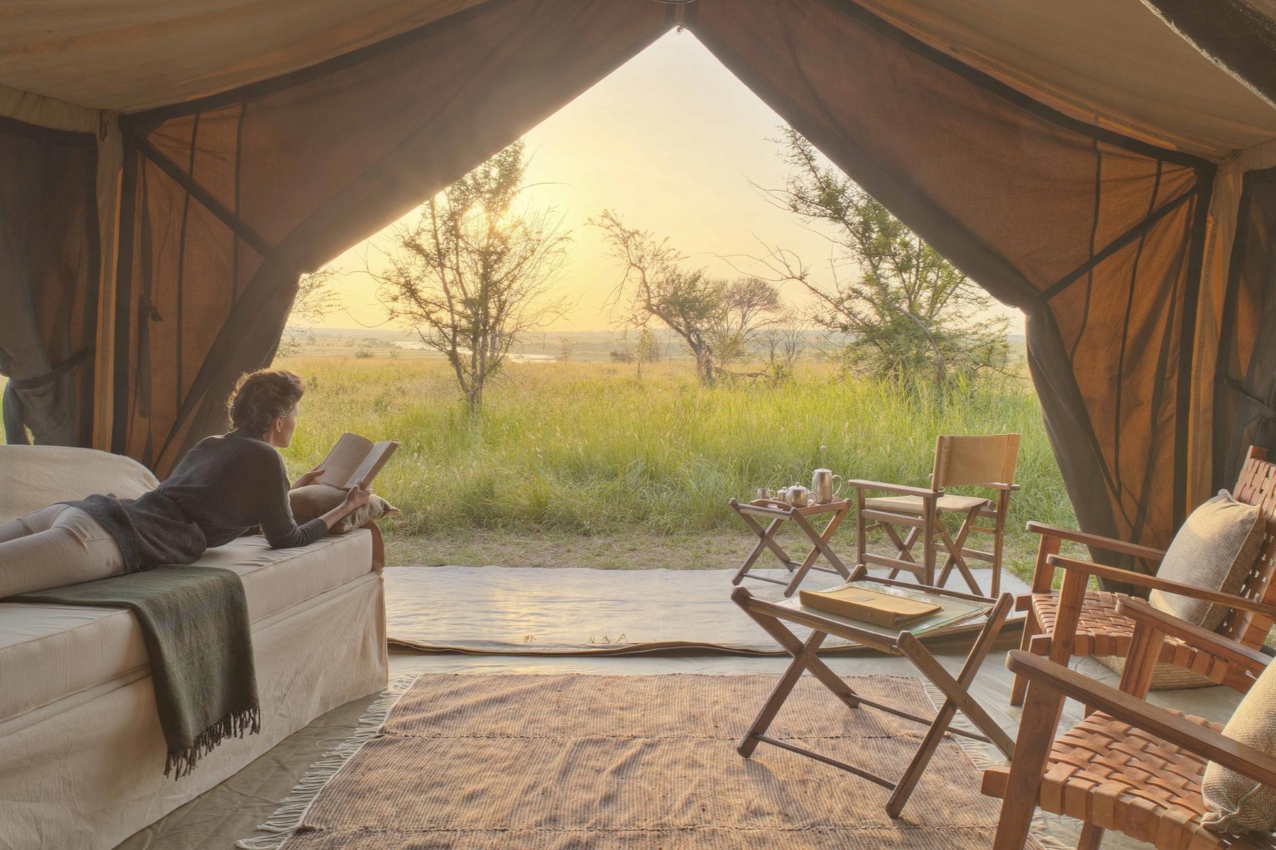 Asilia’s-Olakira-Migration-Camp-cover A woman lounges with a book in one of Africa’s mobile tented camps, facing open canvas flaps and a golden view of the wilderness beyond.