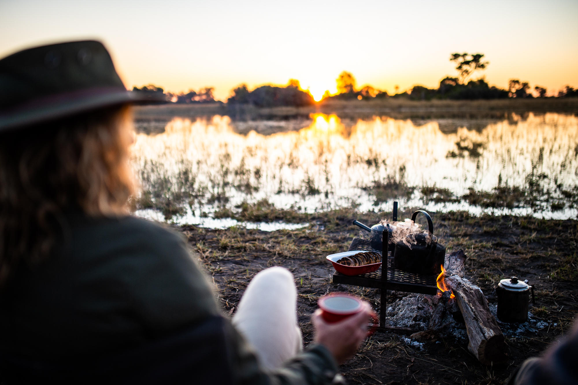 Beagle-Expeditions-Kweene-Trails-Botswana-camp-life A guest at one of Africa’s mobile tented camps sits beside a fire with coffee and rusks at sunrise, overlooking a still floodplain.