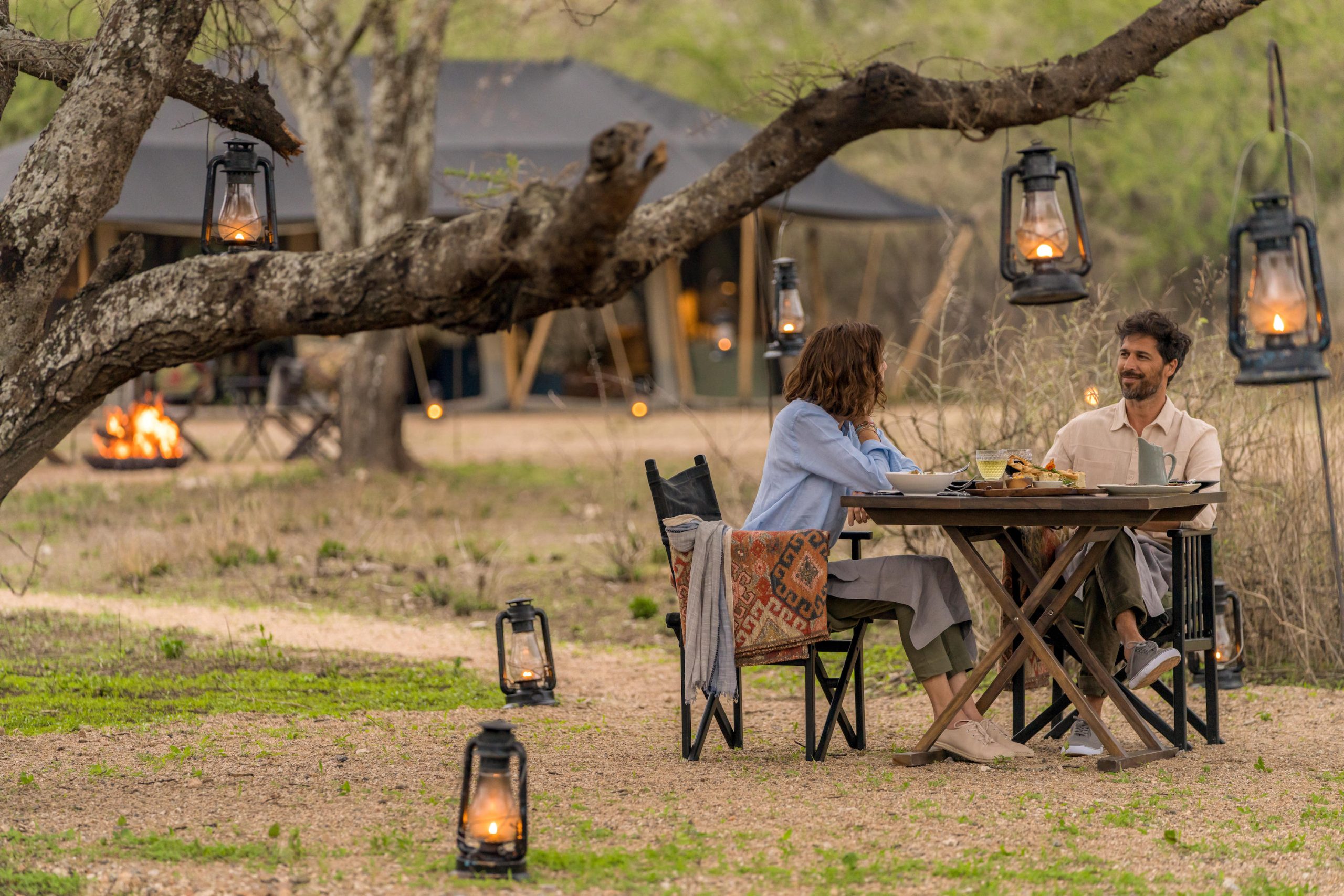 Legendary-Nyasi-Tented-Camp-cover A couple enjoys a lantern-lit meal beneath a tree at one of Africa’s mobile tented camps, with canvas structures and a glowing firepit visible in the background.
