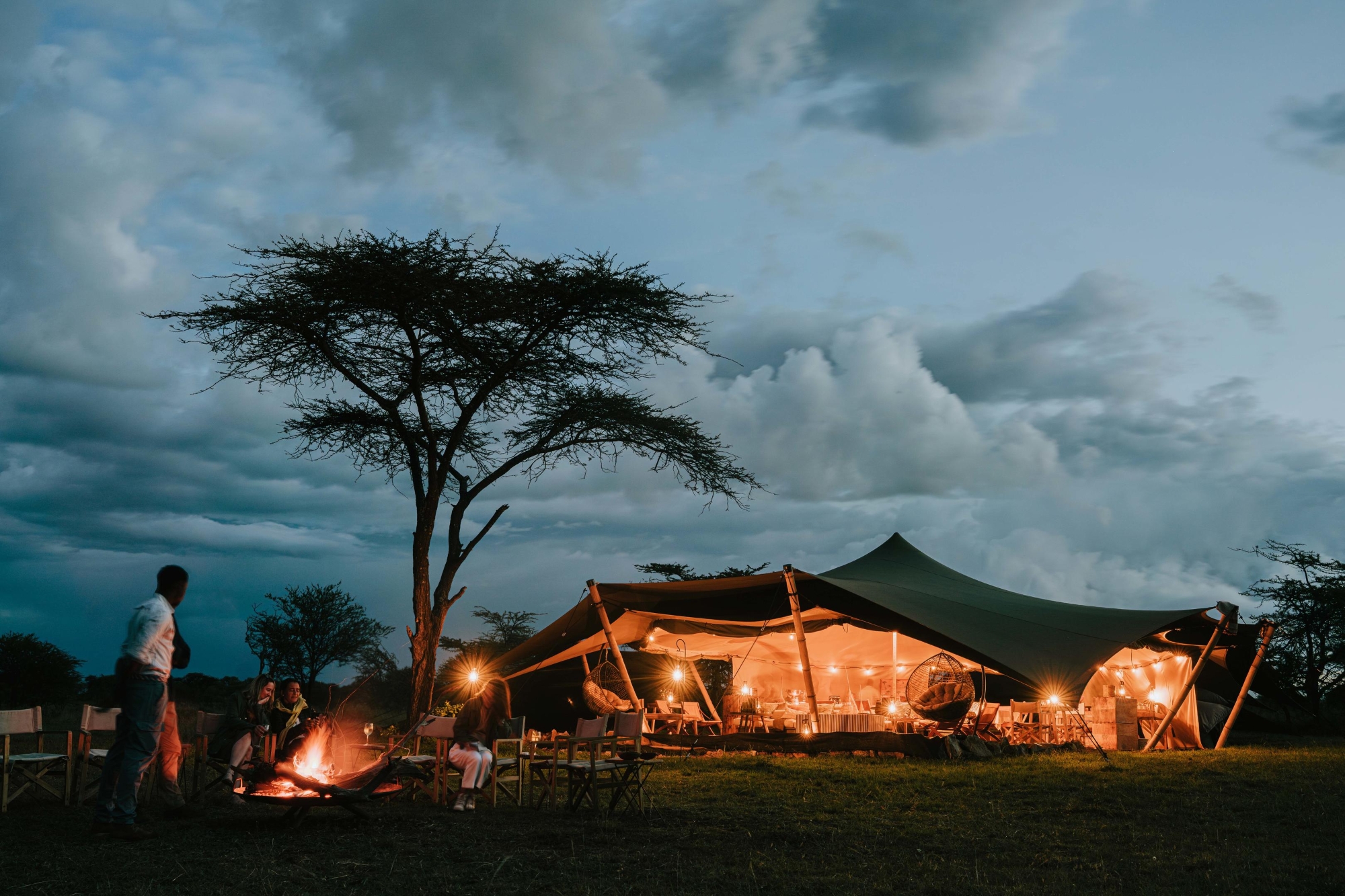 Wilderness-Usawa-Serengeti-campfire A glowing canvas pavilion, lit from within, anchors this mobile tented camp in the Serengeti as guests gather around a fire under the evening sky.