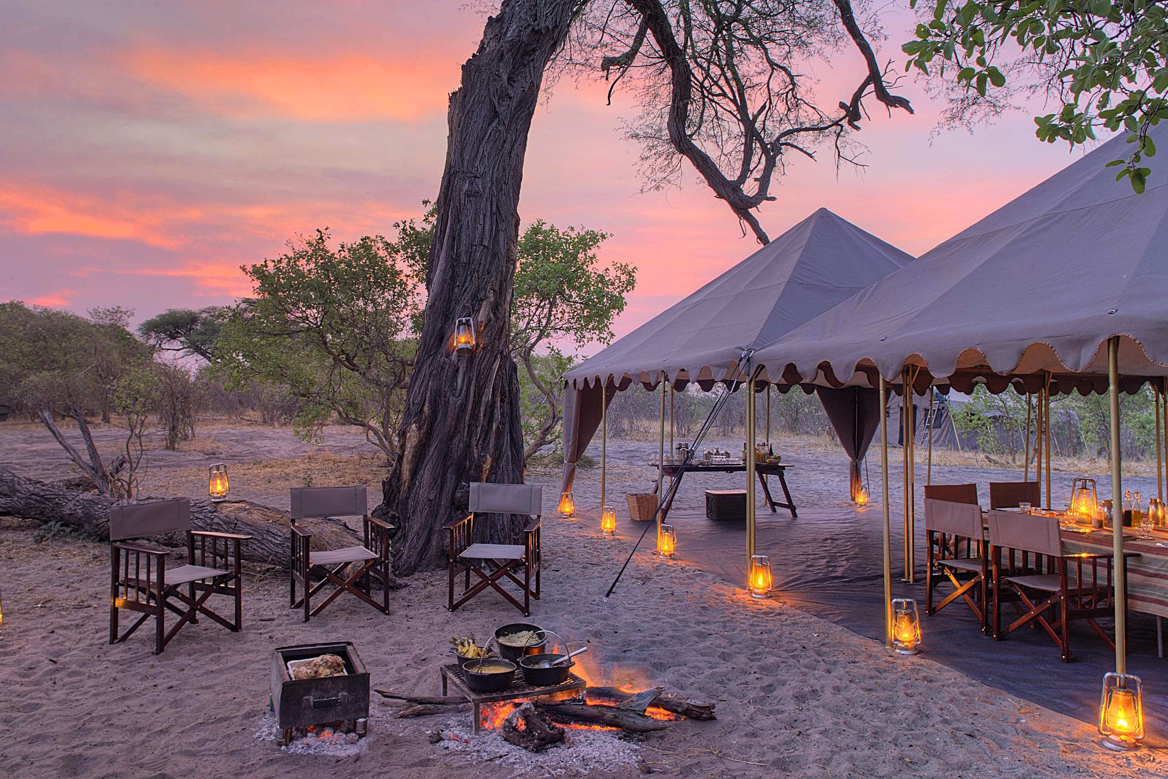 andBeyond-Savute-Under-Canvas-camp-grounds A mobile tented camp glows with lanterns at dusk, featuring canvas dining tents, director's chairs, and a firepit where food is cooked over open coals.