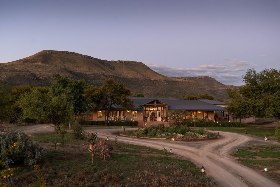 A scenic view of Samara Karoo Lodge nestled at the foot of a mountain, surrounded by natural vegetation at dusk.