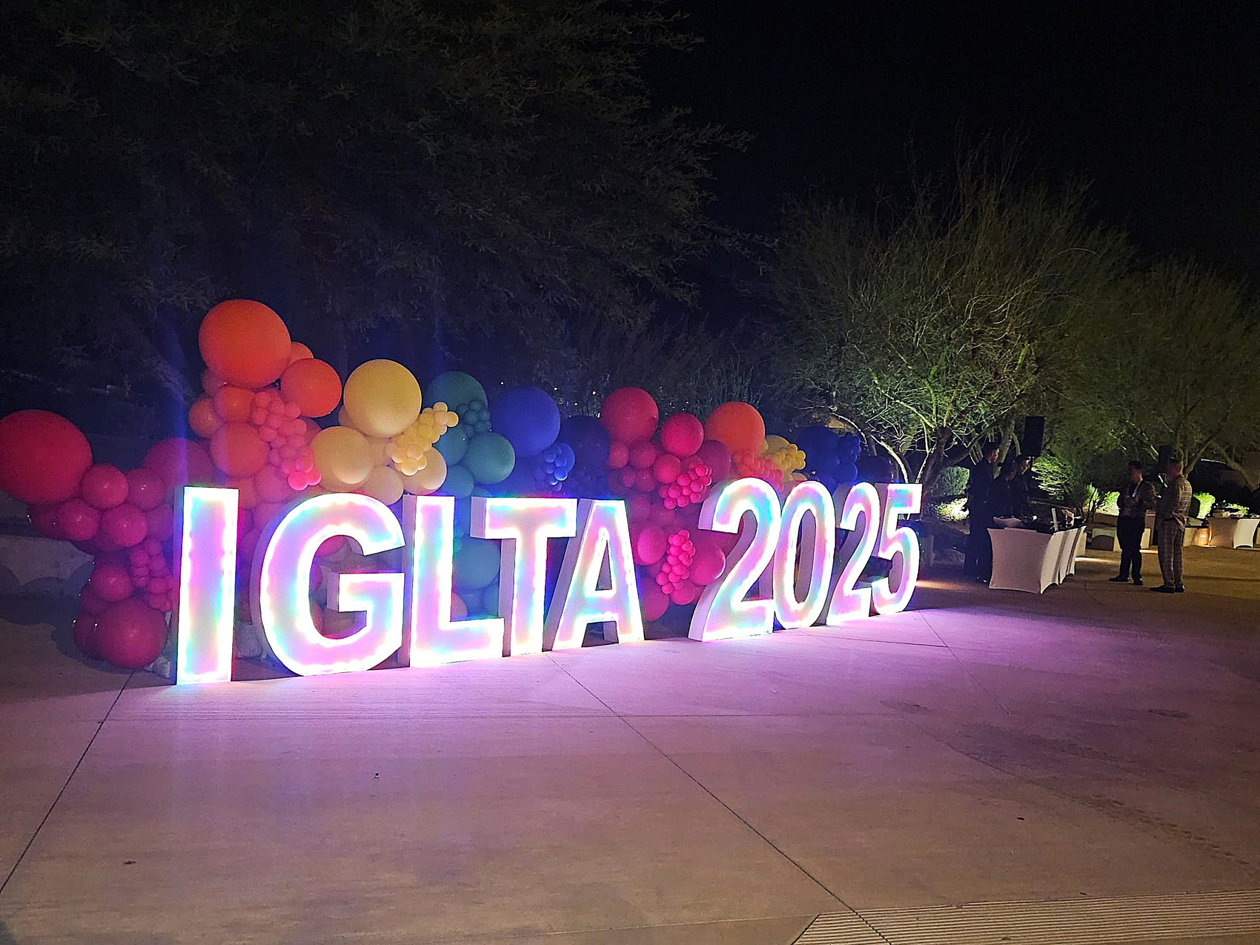 Large illuminated letters spelling “IGLTA 2025” stand against a backdrop of rainbow-coloured balloons at an outdoor night event.