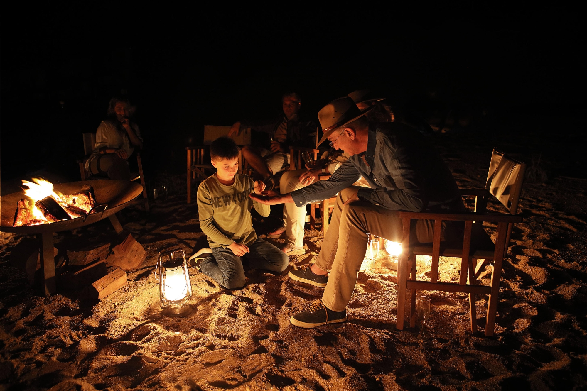 Londolozi-Varty-Camp-campfire-story A group gathers around a fire at night as an adult leans toward a child in a warm, guiding gesture, a small scene echoing how Londolozi’s legacy has always passed from one generation to the next.