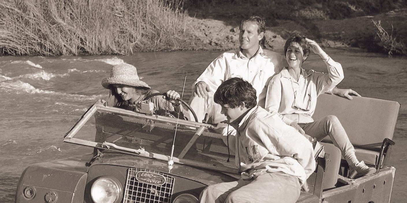 Four people sit in an old Land Rover and cross a river while on safari