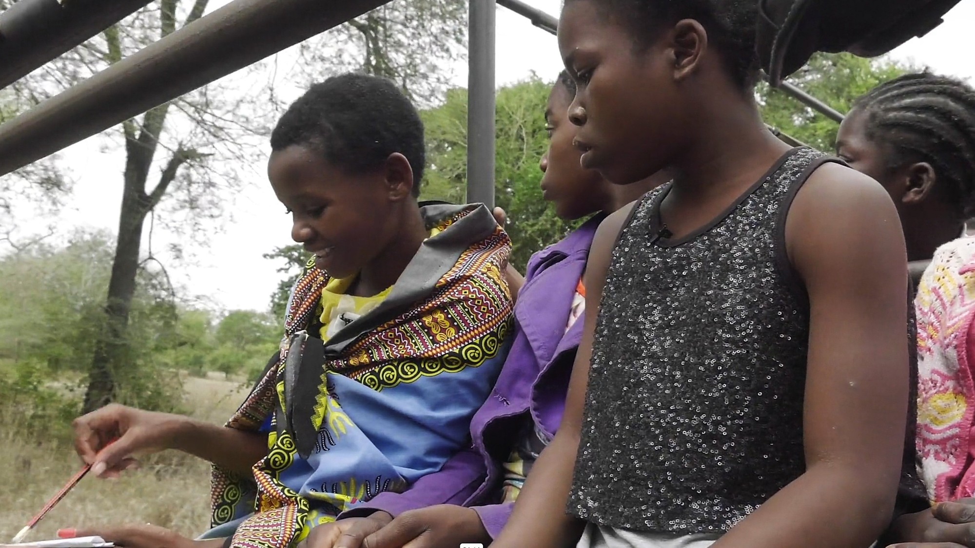 Sam-gorongosa-safari-girls-club Girls sit together on a vehicle bench, one drawing in her notebook during a Gorongosa safari.