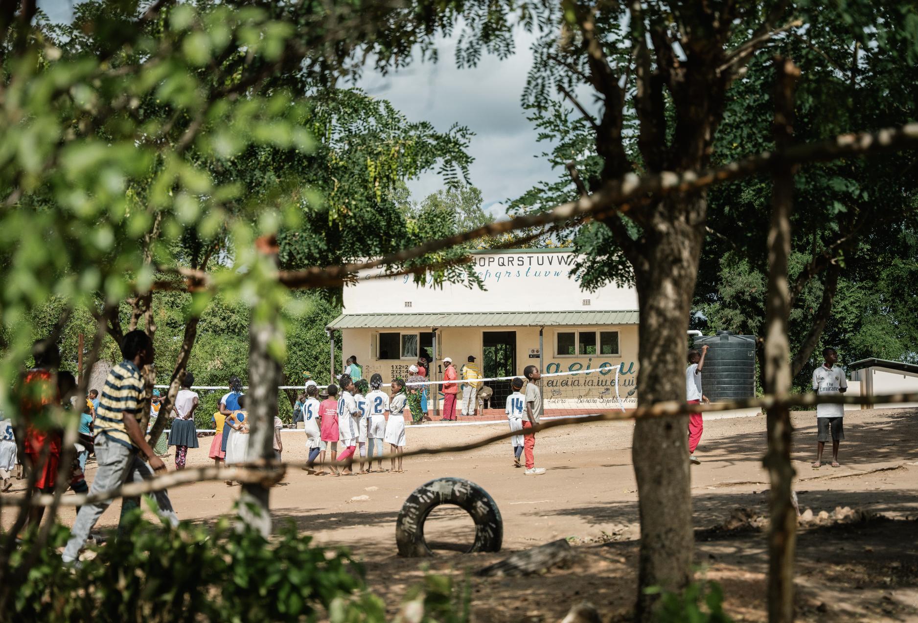 1752754694851_Gorongosa-Safaris---Community-Visits1 Children gather and play outside a rural school building shaded by trees, showing how education and everyday life are strengthened through the impact in Gorongosa.