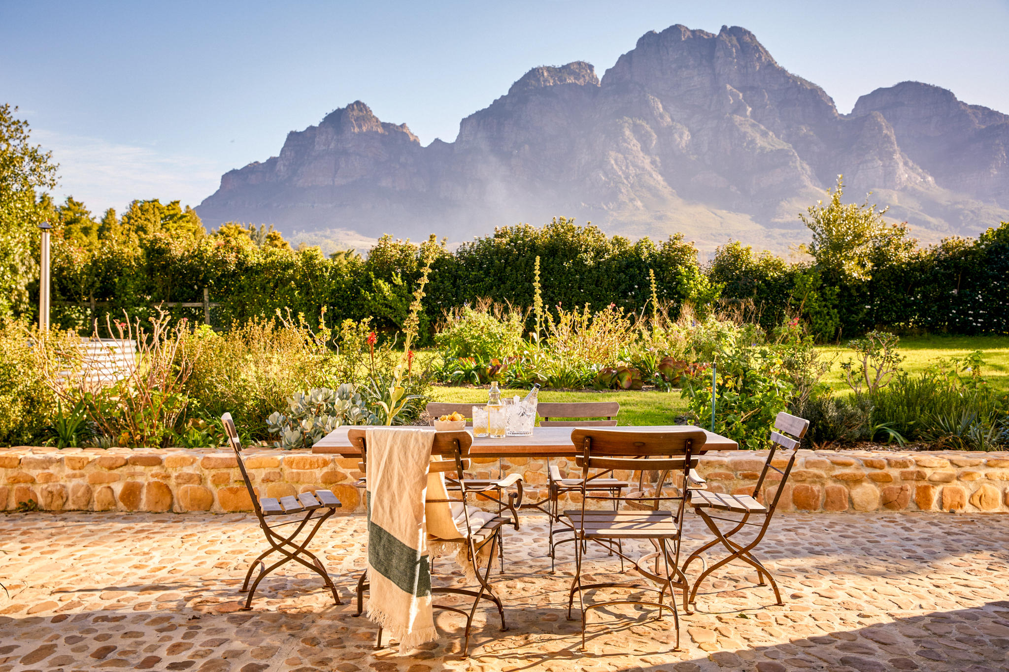 An outdoor wooden dining table set for two sits on a sun-warmed stone terrace, framed by lush gardens and dramatic mountain peaks, capturing the quiet romance of a South African honeymoon.