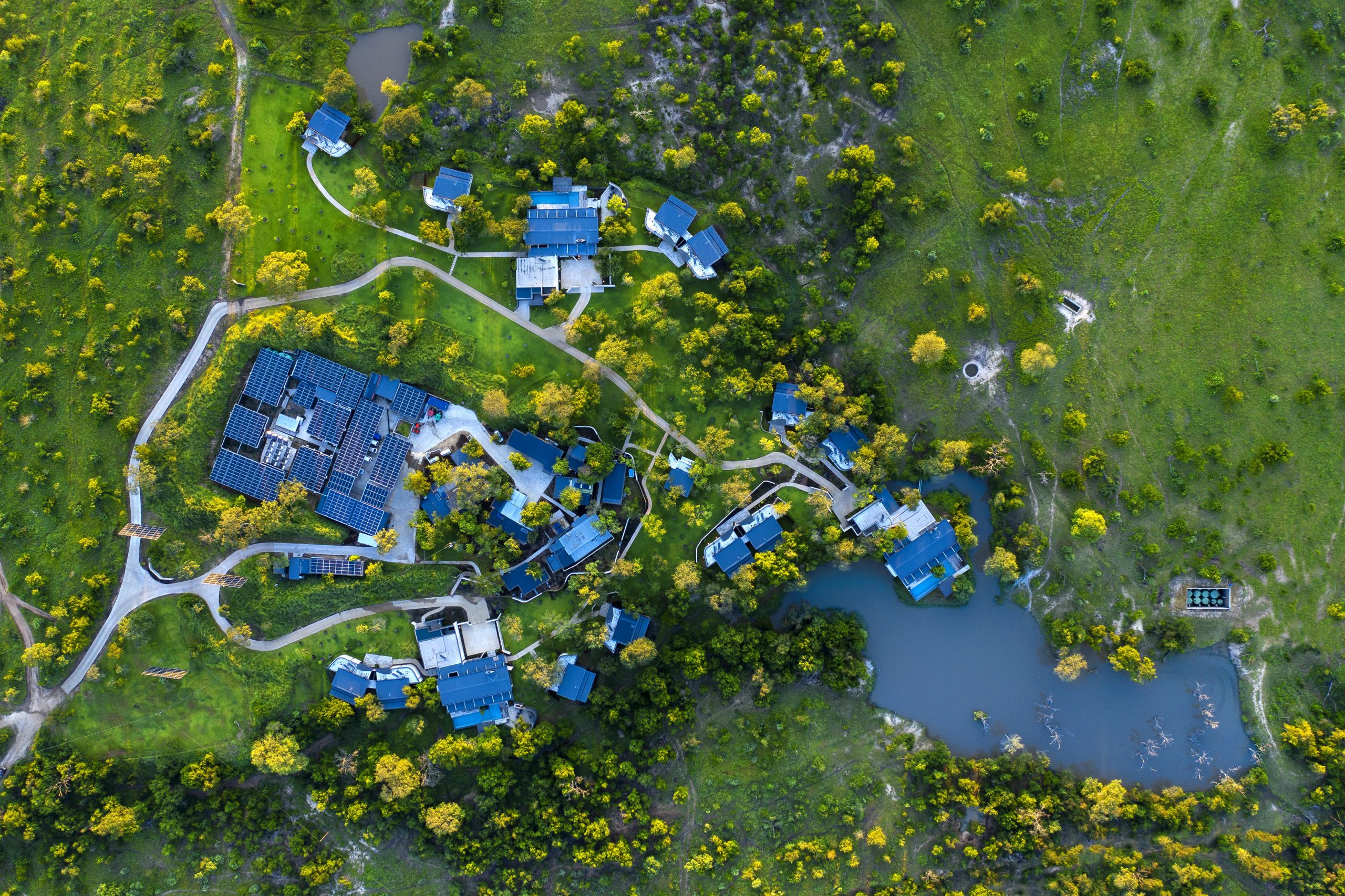 An aerial view of a modern safari lodge spread across green bushveld, with villas clustered around a prominent waterhole, revealing how lodges with the best waterholes are thoughtfully shaped by water and space.