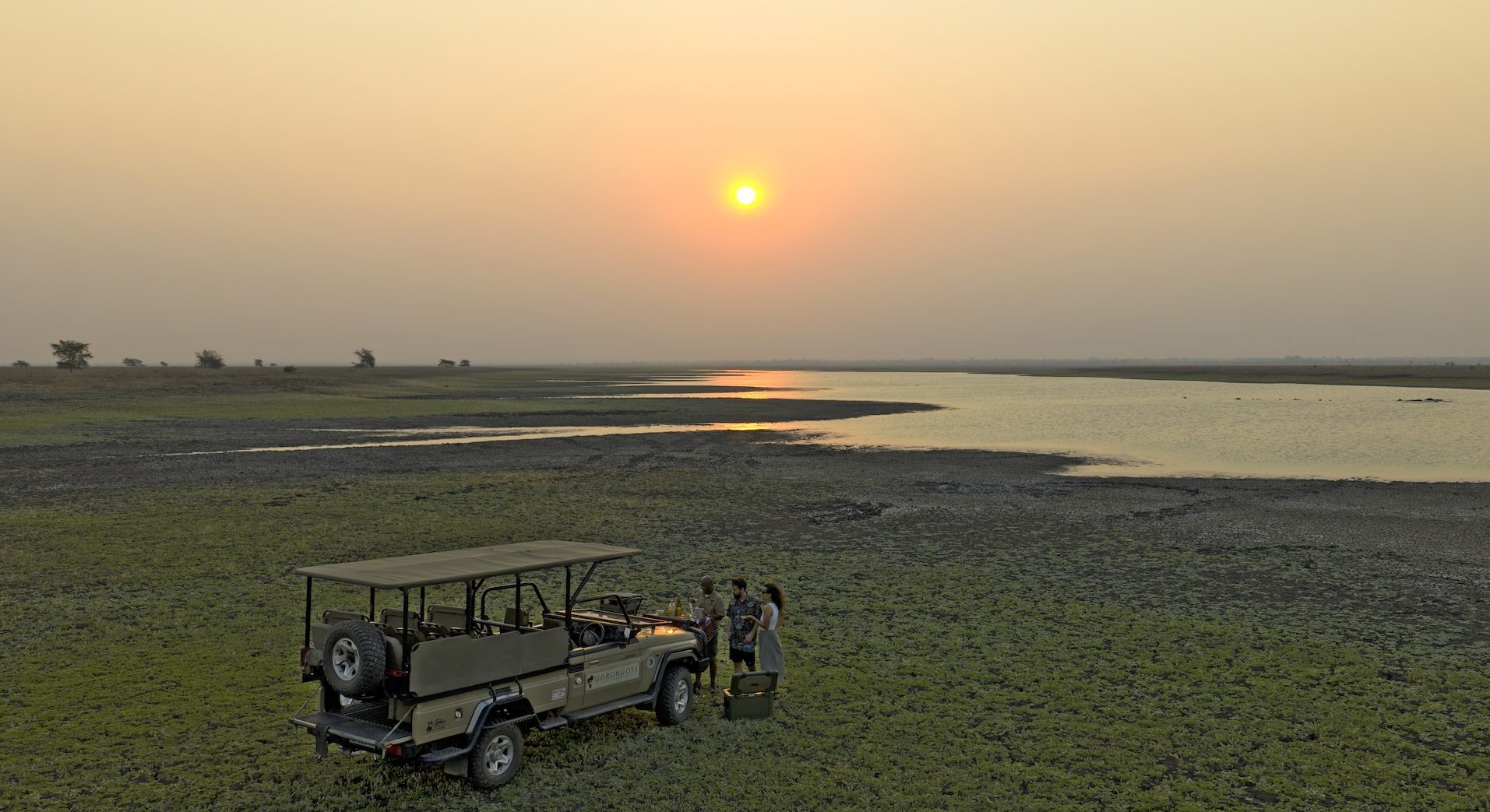 Guests pause beside a safari vehicle at sunset overlooking a winding waterway, capturing a moment of quiet connection shaped by the impact in Gorongosa.