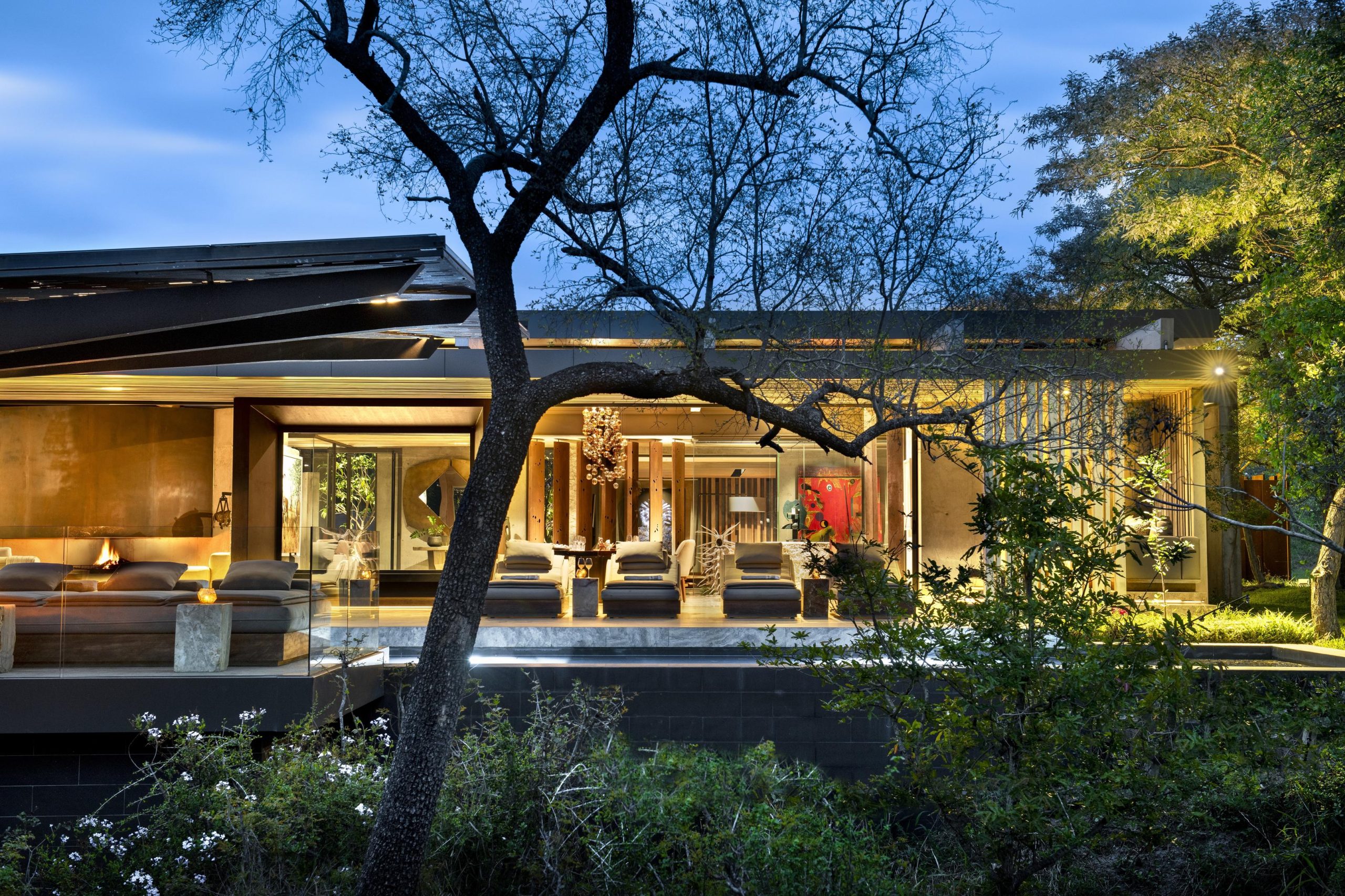 A softly lit, modern safari lodge glowing at dusk among trees beside a nearby water source, capturing the quiet elegance of lodges with the best waterholes.
