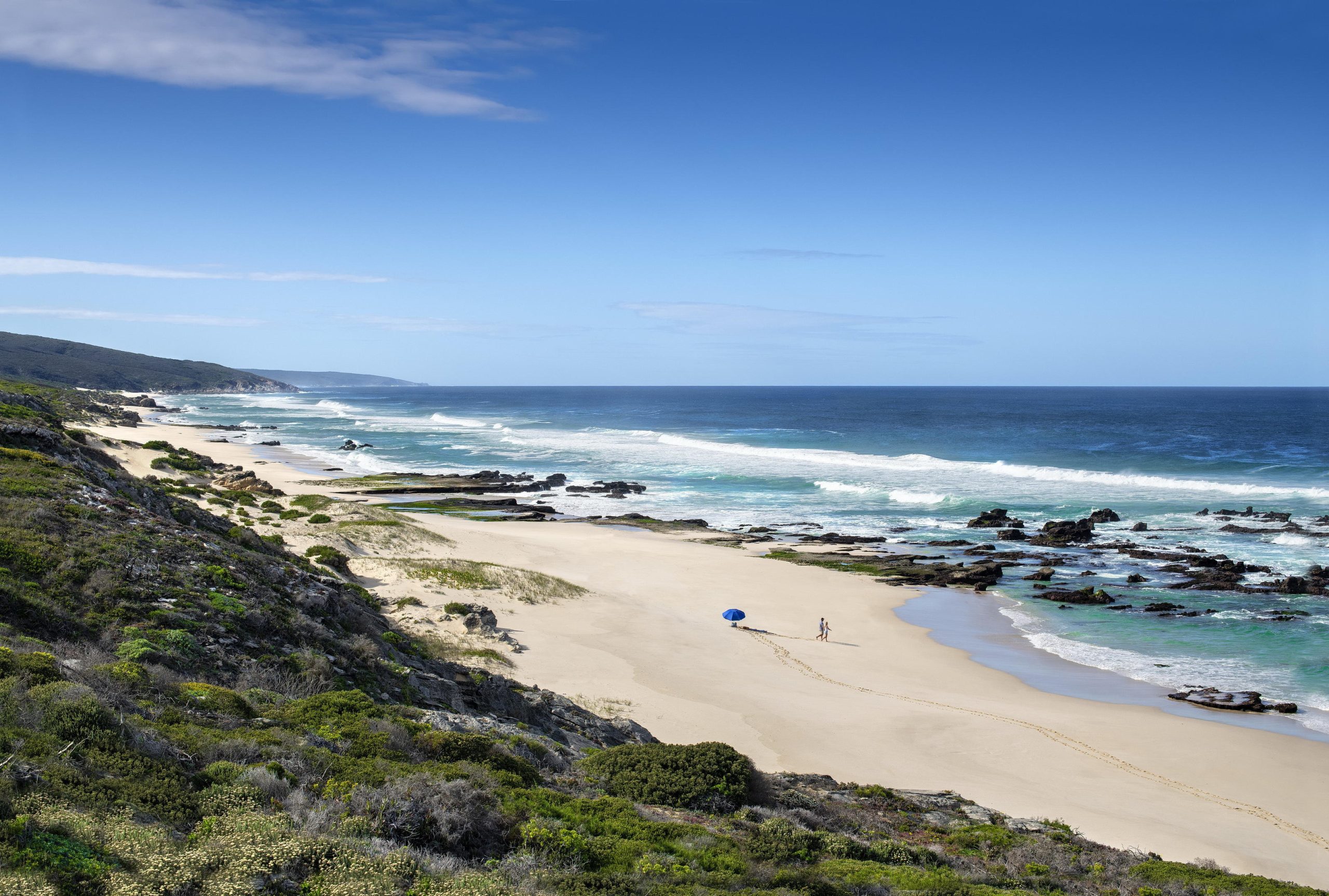 A vast, empty stretch of pale sand curves along a wild coastline, with rolling waves, rocky outcrops, and two tiny figures walking beneath a wide blue sky on a South African honeymoon.