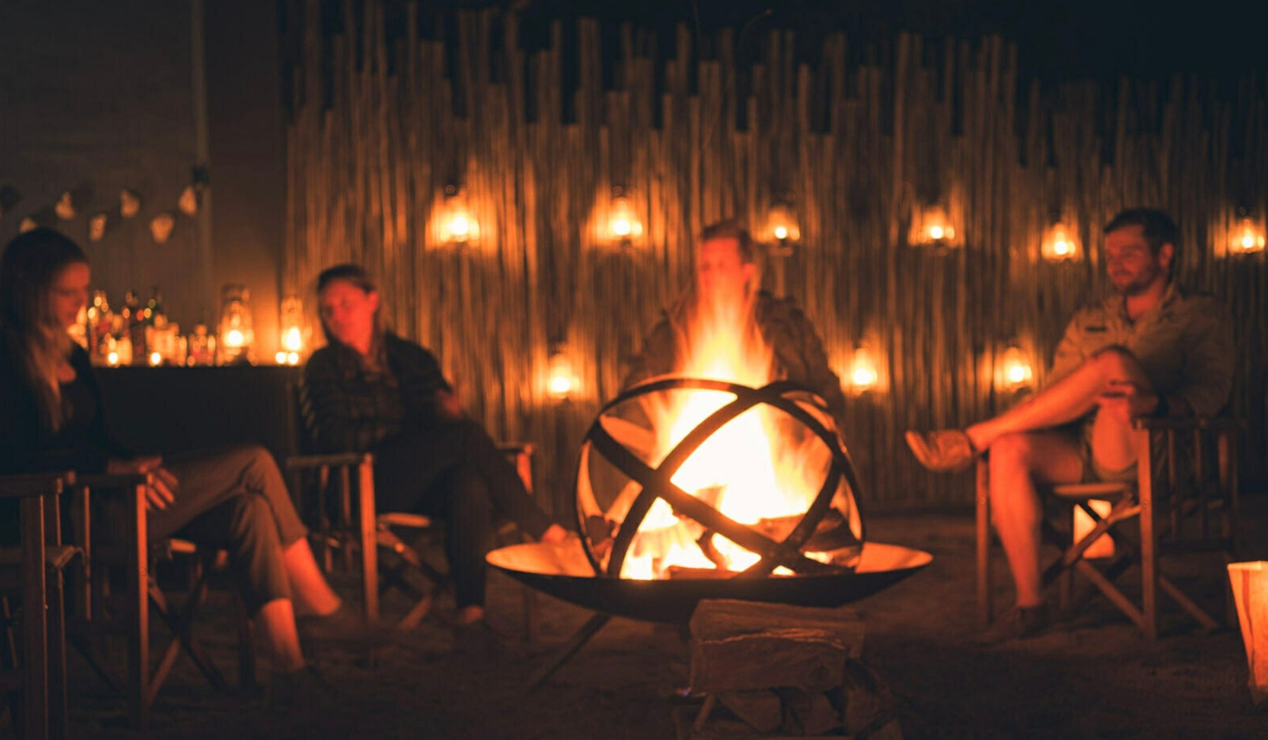 A small group of people sit in wooden chairs around a glowing firepit at night, lanterns casting warm light, reflecting the shared, unguarded moments that define Rhino Africa and Londolozi.