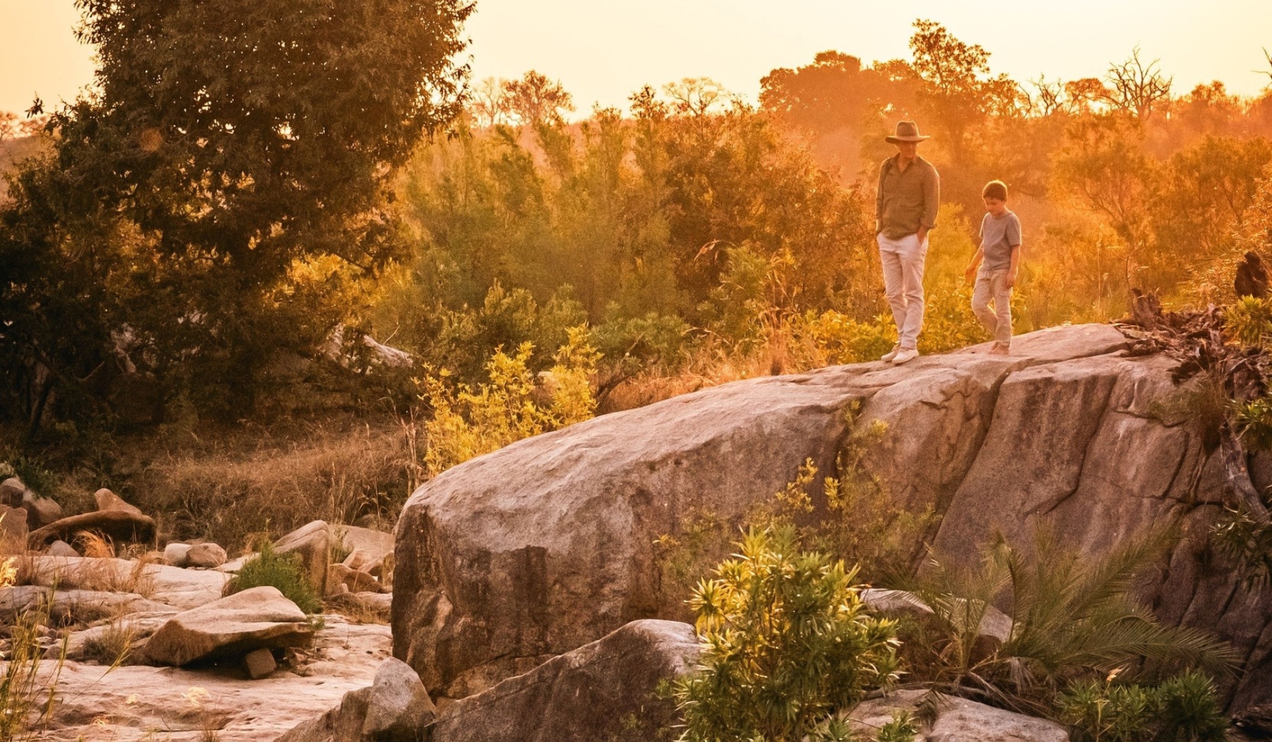 An adult and a child walk carefully across a large rock at golden hour, surrounded by bushveld, reflecting the shared values and generational thinking that connect Rhino Africa and Londolozi.
