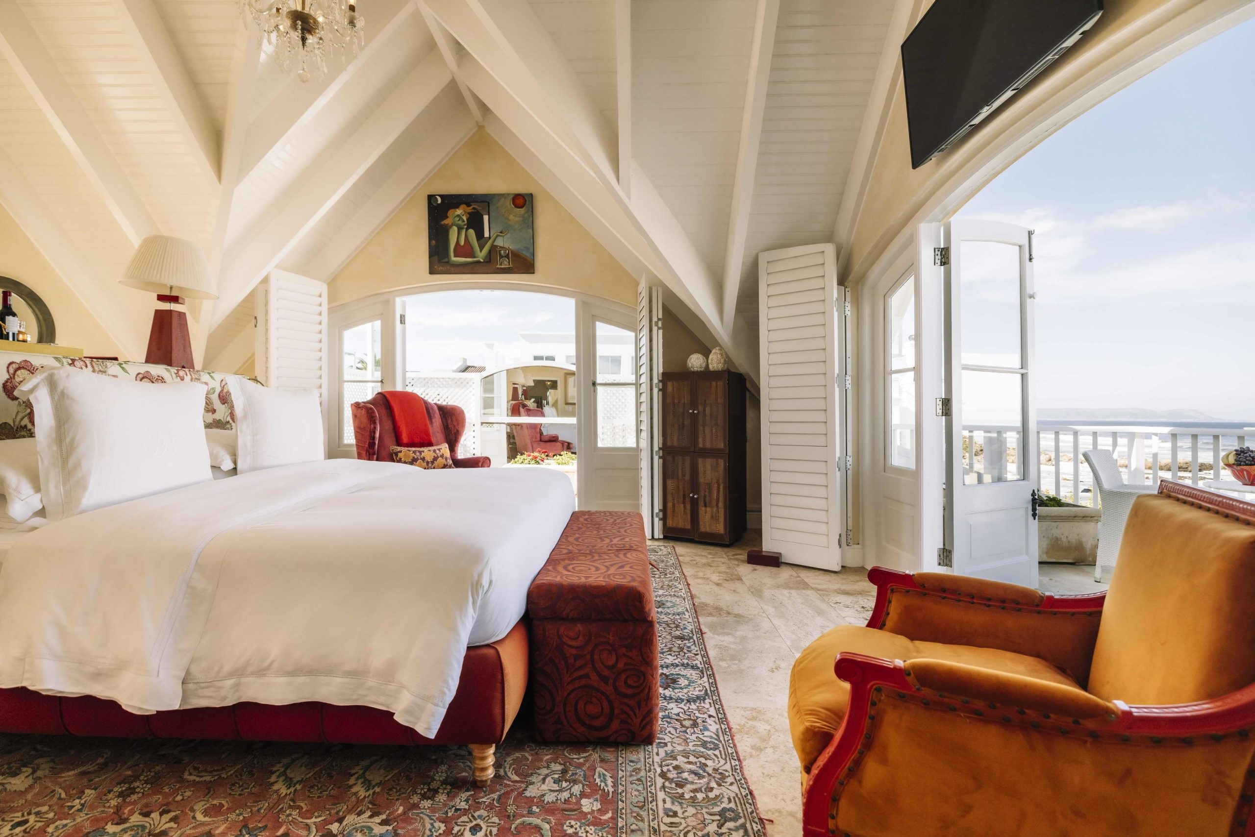 A light-filled attic-style bedroom with crisp white linens, warm armchairs, and open doors leading to a sea-view balcony evokes the comfort and indulgence of a South African honeymoon.