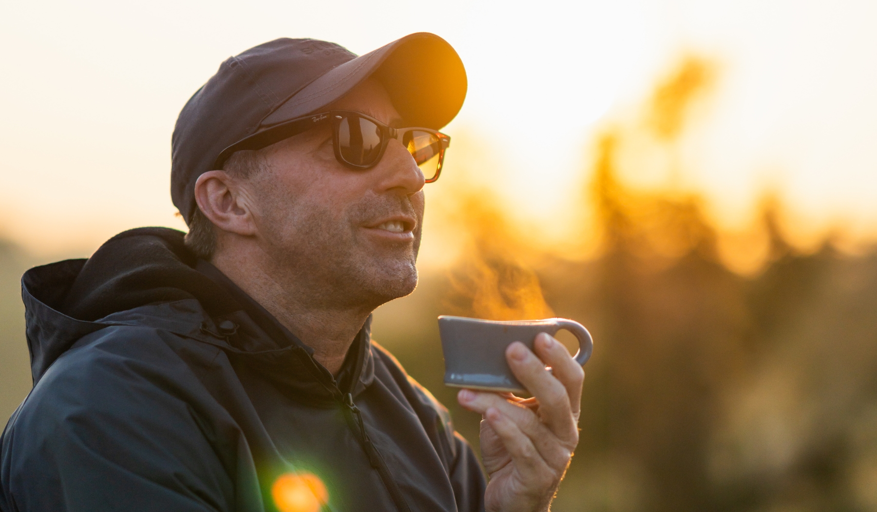 David Ryan in a cap and sunglasses holds a steaming cup outdoors at sunrise, pausing in quiet thought during a reflective moment linked to Rhino Africa and Londolozi.