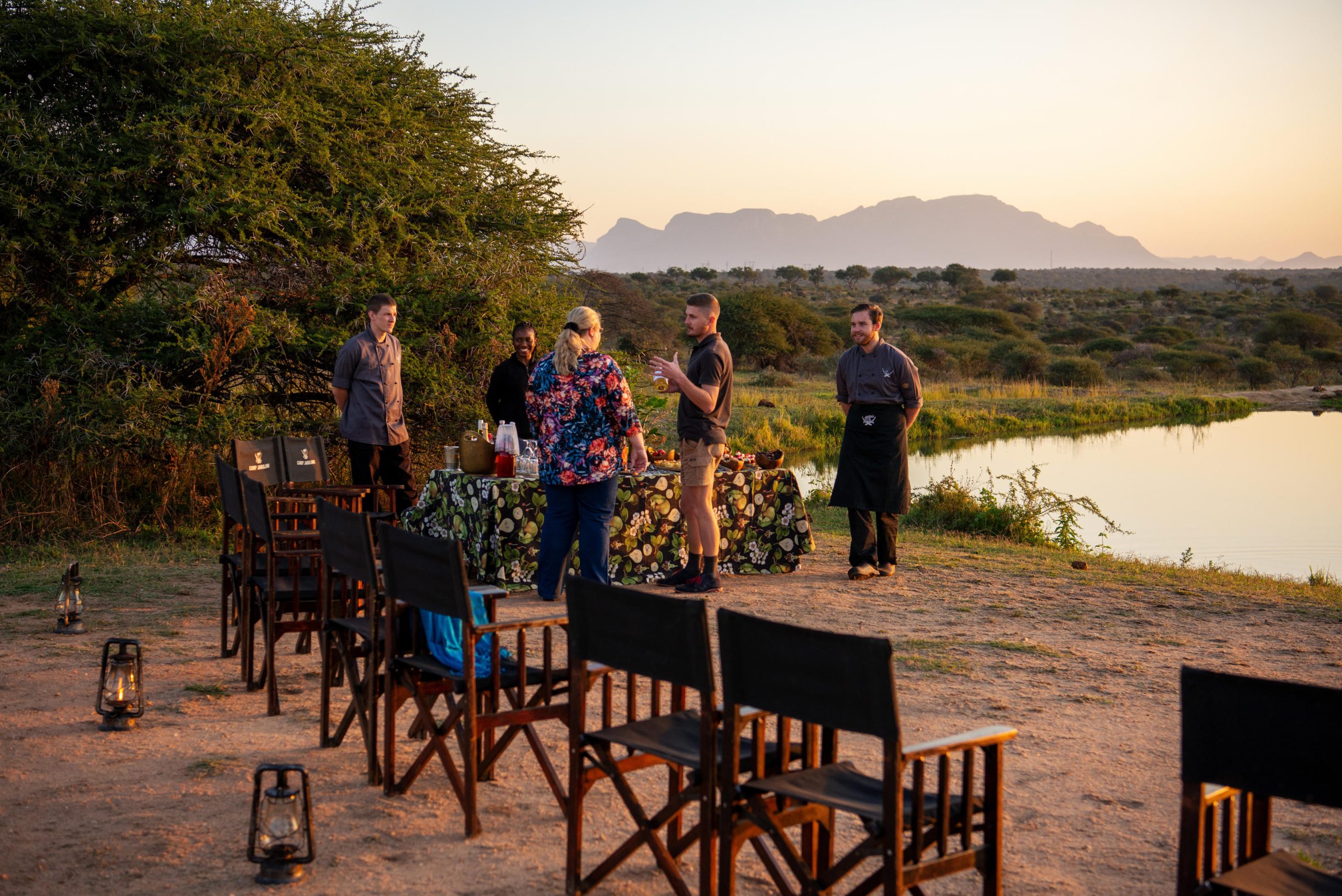 A safari team prepares an elegant sundowner setup beside a still waterhole at sunset, reflecting the intimate, unhurried moments offered by lodges with the best waterholes.
