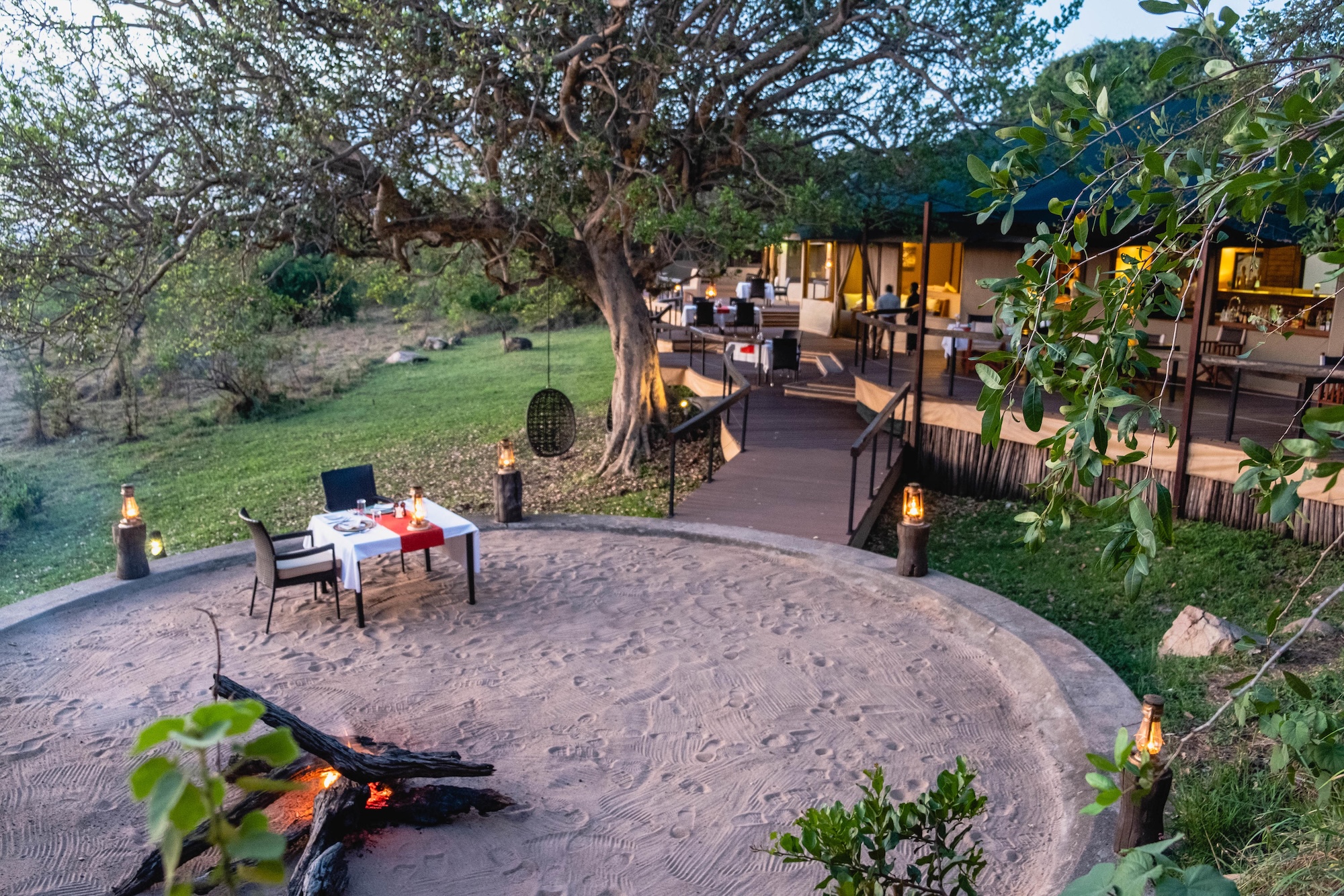 A candlelit dinner beside a small fire pit sits beneath a sprawling tree outside a tented suite, capturing the intimate atmosphere found at Luxury safari lodges in Tanzania.