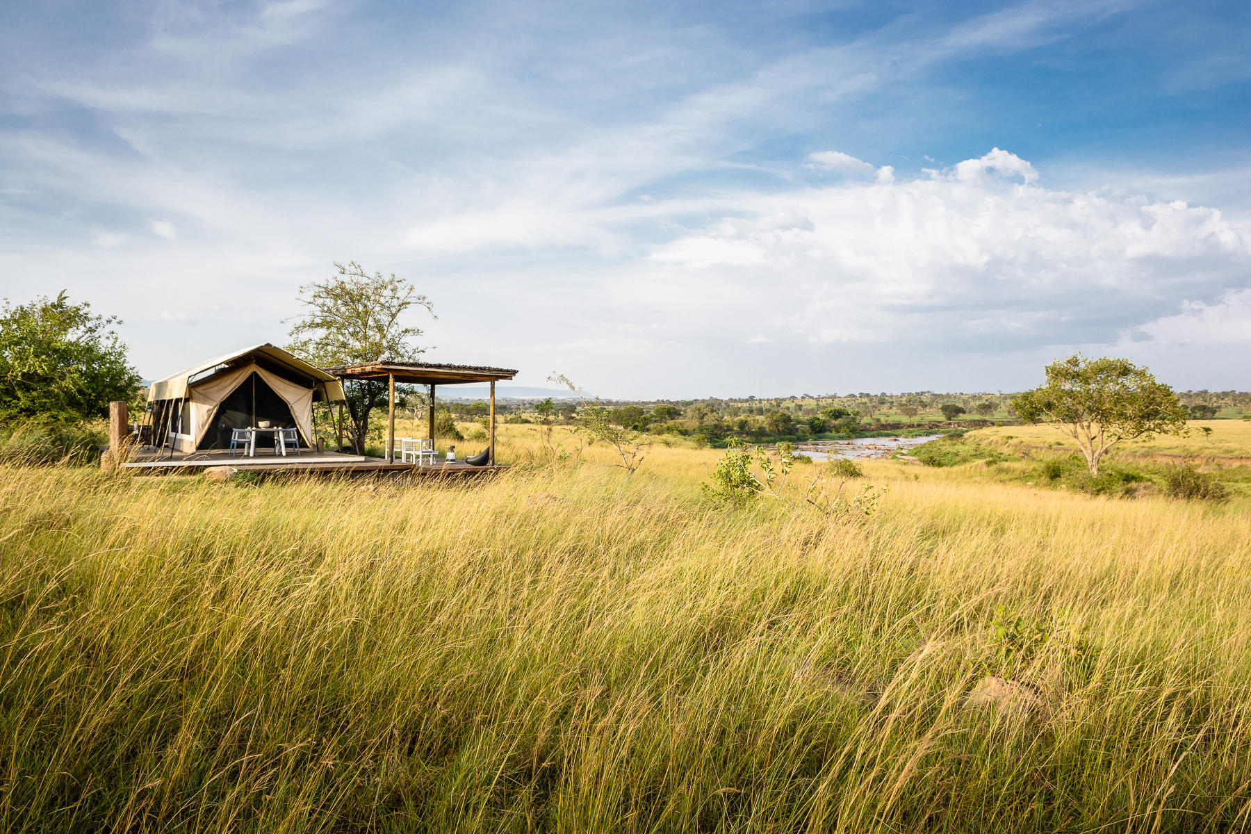 A solitary canvas tent on a raised wooden deck overlooks golden grasslands and a winding river, embodying the remote intimacy of Luxury safari lodges in Tanzania.