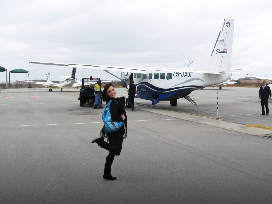 A Rhino Africa travel consultant with a blue backpack smiles on the tarmac beside a small propeller plane as staff prepare for boarding.