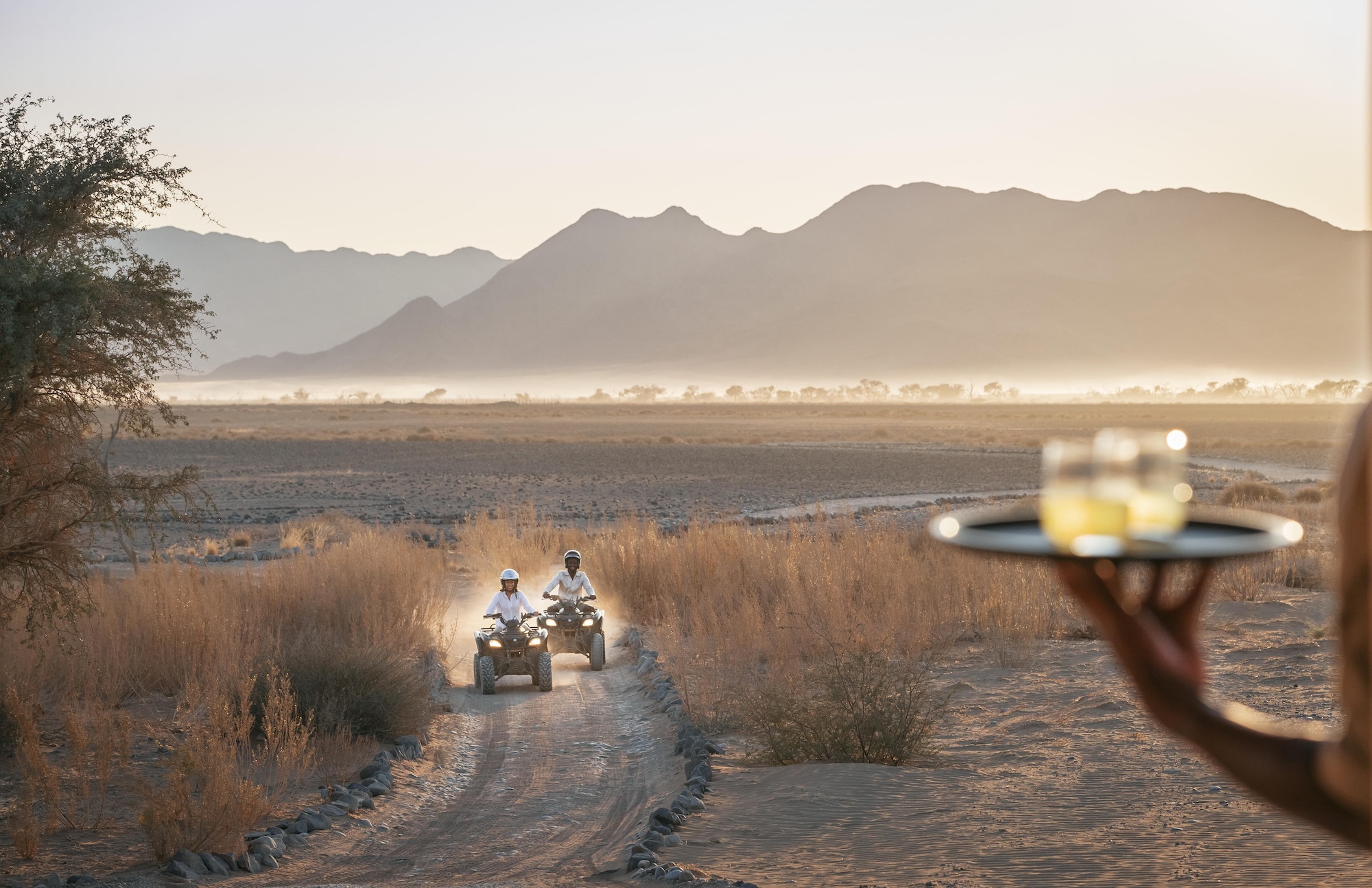 Two travellers ride quad bikes through a dry riverbed at Little Kulala, with soft desert light and mountains rising in the distance.