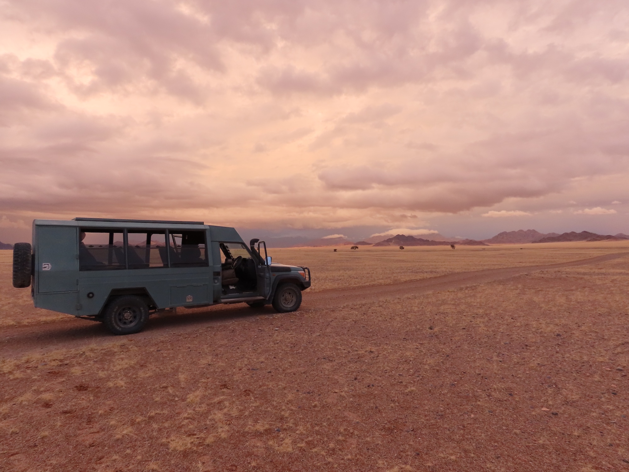 Safari vehicle parked on an open Namibian plain beneath a vast pink-grey sky at sunrise.