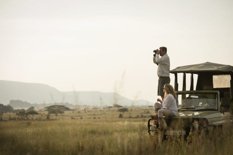 Guests on a Dunia Camp game drive vehicle in Tanzania’s Serengeti, with one person standing to photograph the open savannah while another sits on the bonnet overlooking the plains.