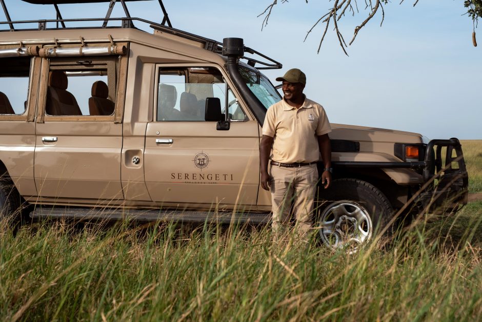 A Serengeti Sojourn Camp guide standing beside a safari vehicle in the grassy plains of Tanzania’s Serengeti.